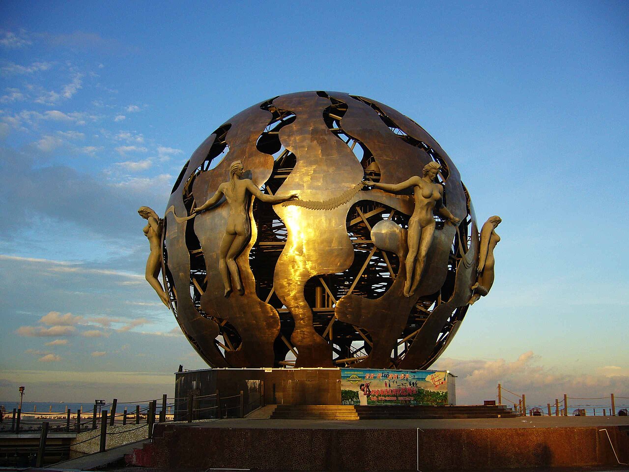 a large openwork metal sphere with figures of women around it against a blue evening sky