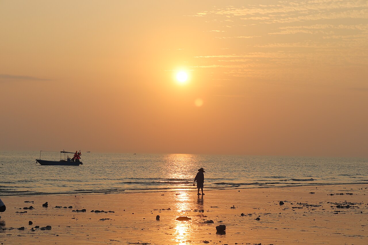 the sun low over the sea at a rocky beach with a small boat and a figure at the water's edge