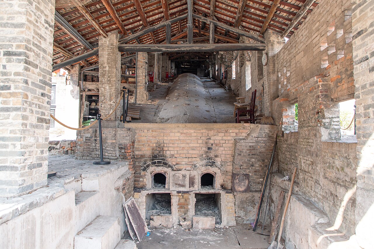 inside a long brick kiln shed with wooden roof beams and arched kiln openings at floor level