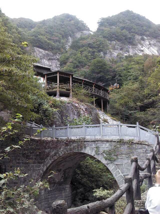 Stone arch bridge and wooden hillside building below forested cliffs in the Dabie Mountains