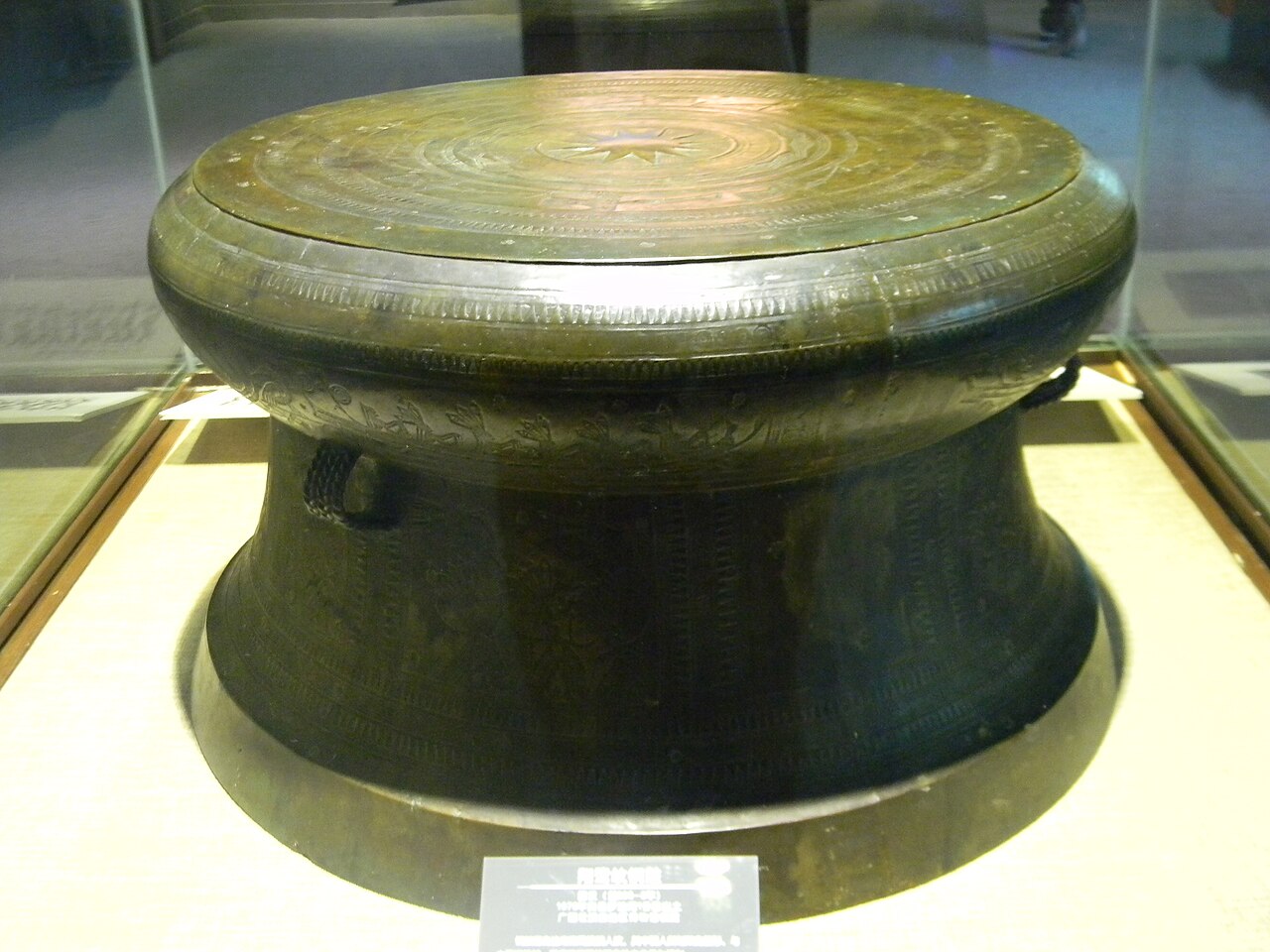 a large dark bronze drum with a flat decorated top in a lit display case