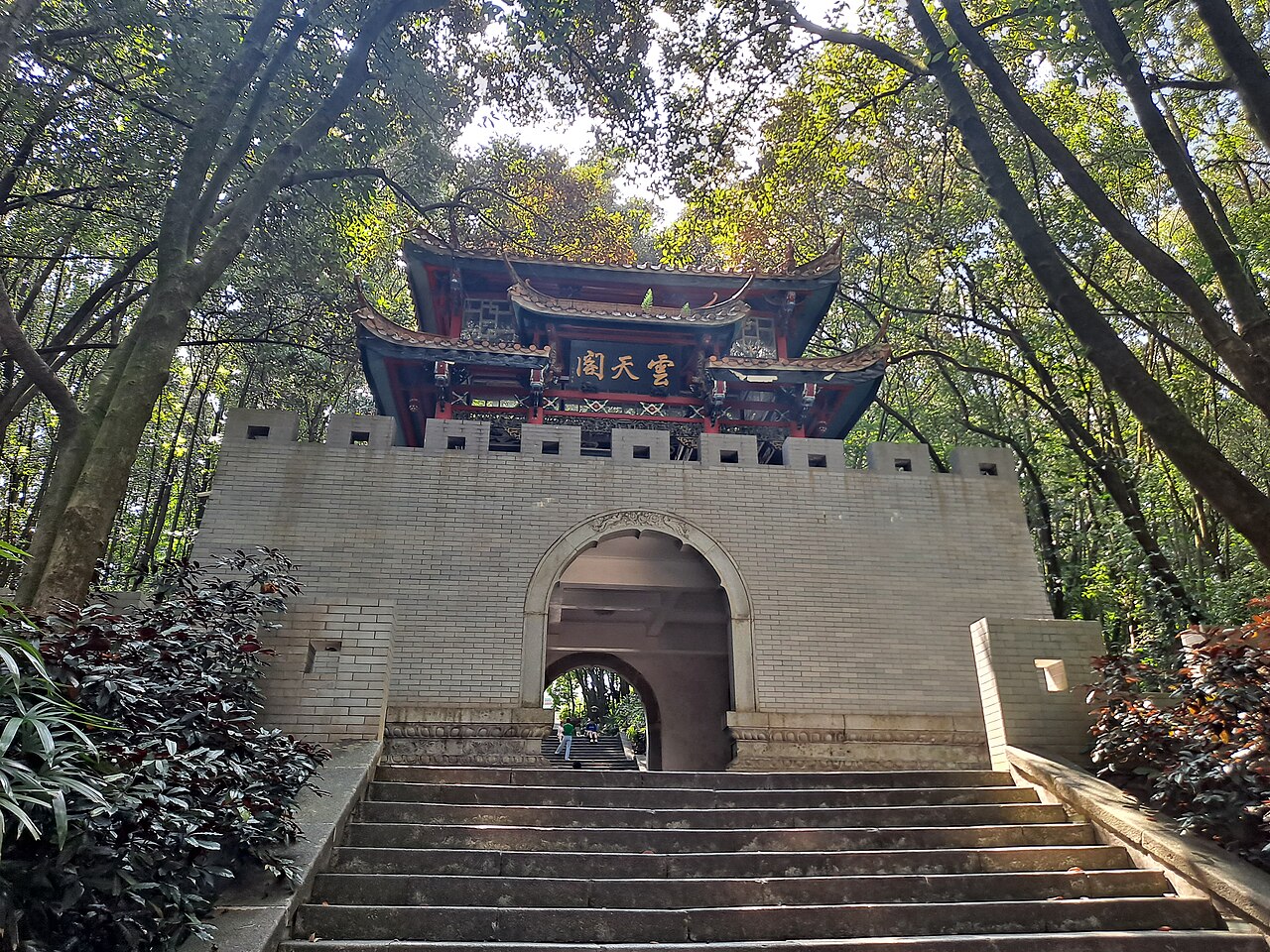 a stone gate with a red and gold pavilion on top at the head of a flight of steps among trees