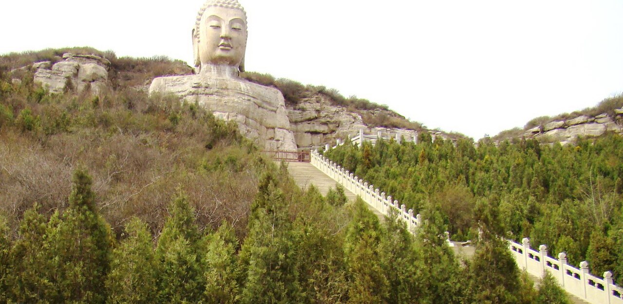 Giant stone Buddha head and shoulders rising above pine woods and a long stairway on Mengshan
