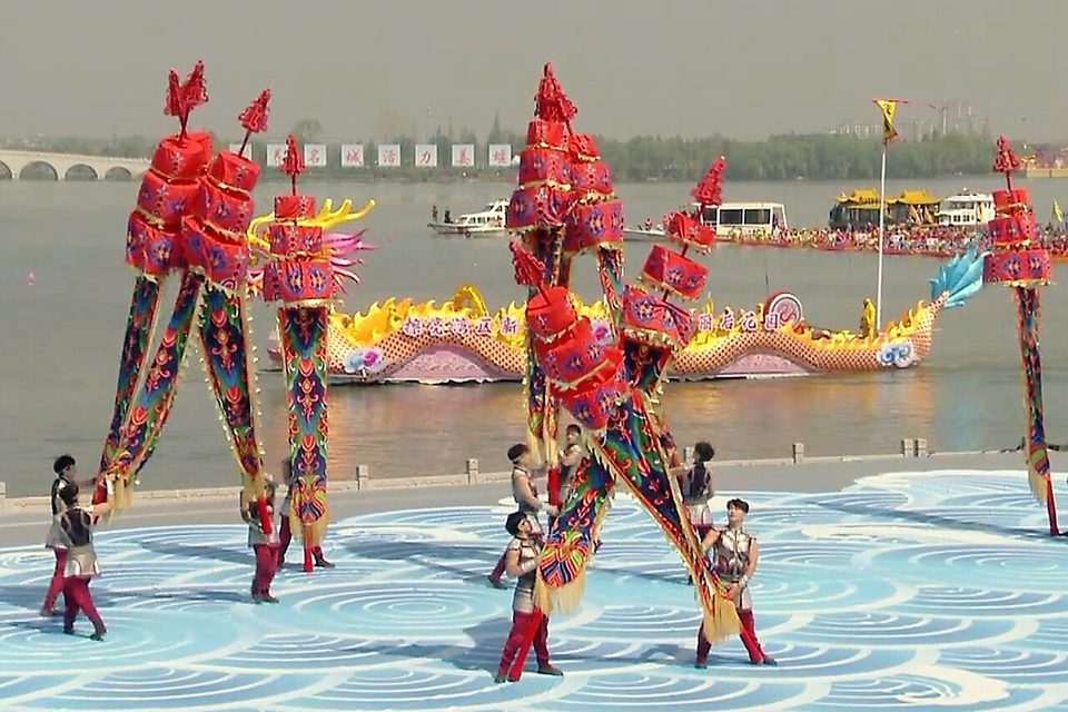 Performers in red costumes with tall decorations at the Qintong Boat Festival, with boats on the lake behind