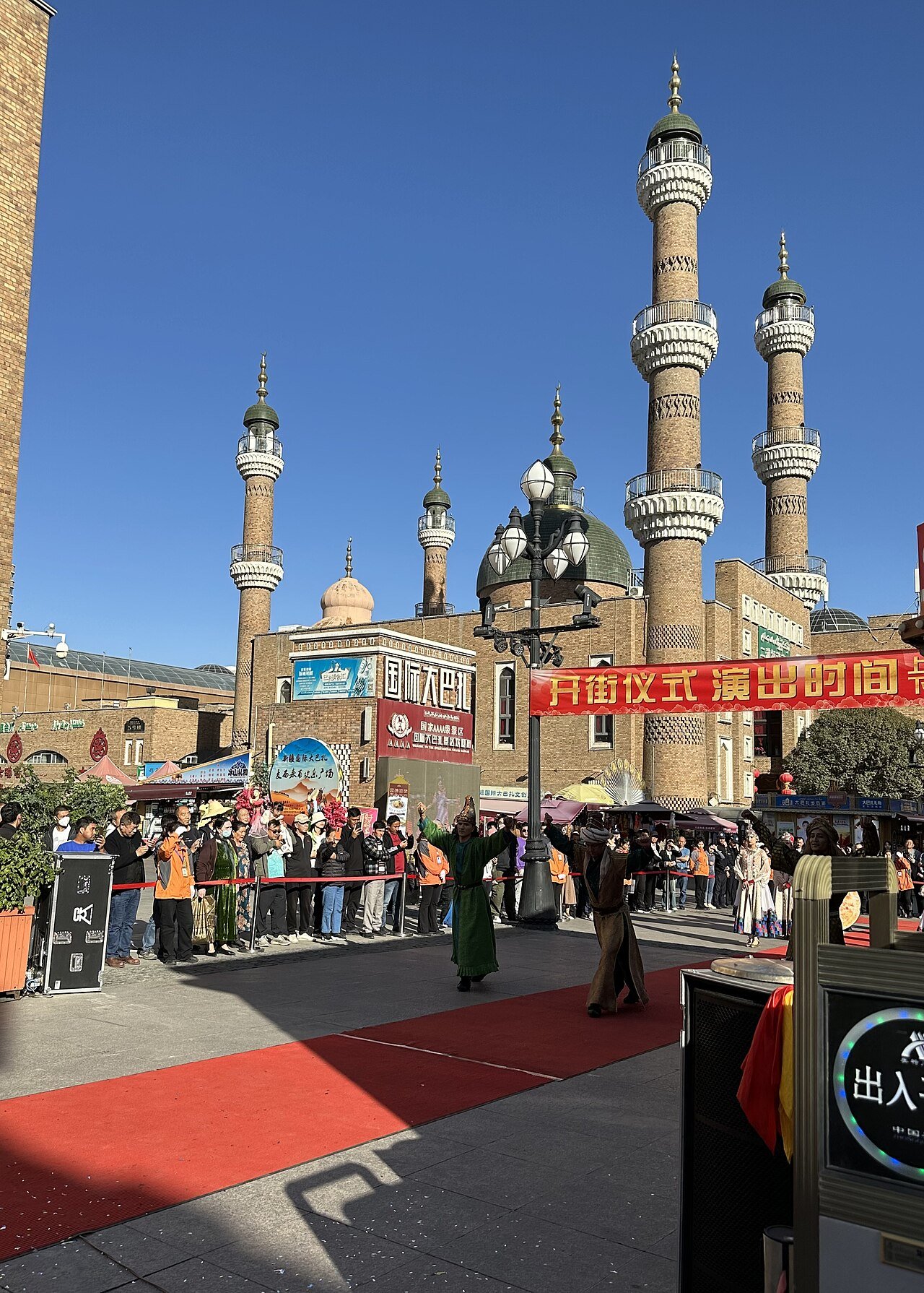 performers in costume on a red carpet in the bazaar pedestrian street with minarets behind and a crowd watching