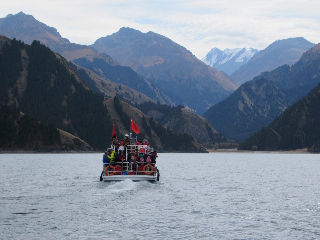 a red tour boat crossing the grey-blue lake toward forested mountains and a snowy peak
