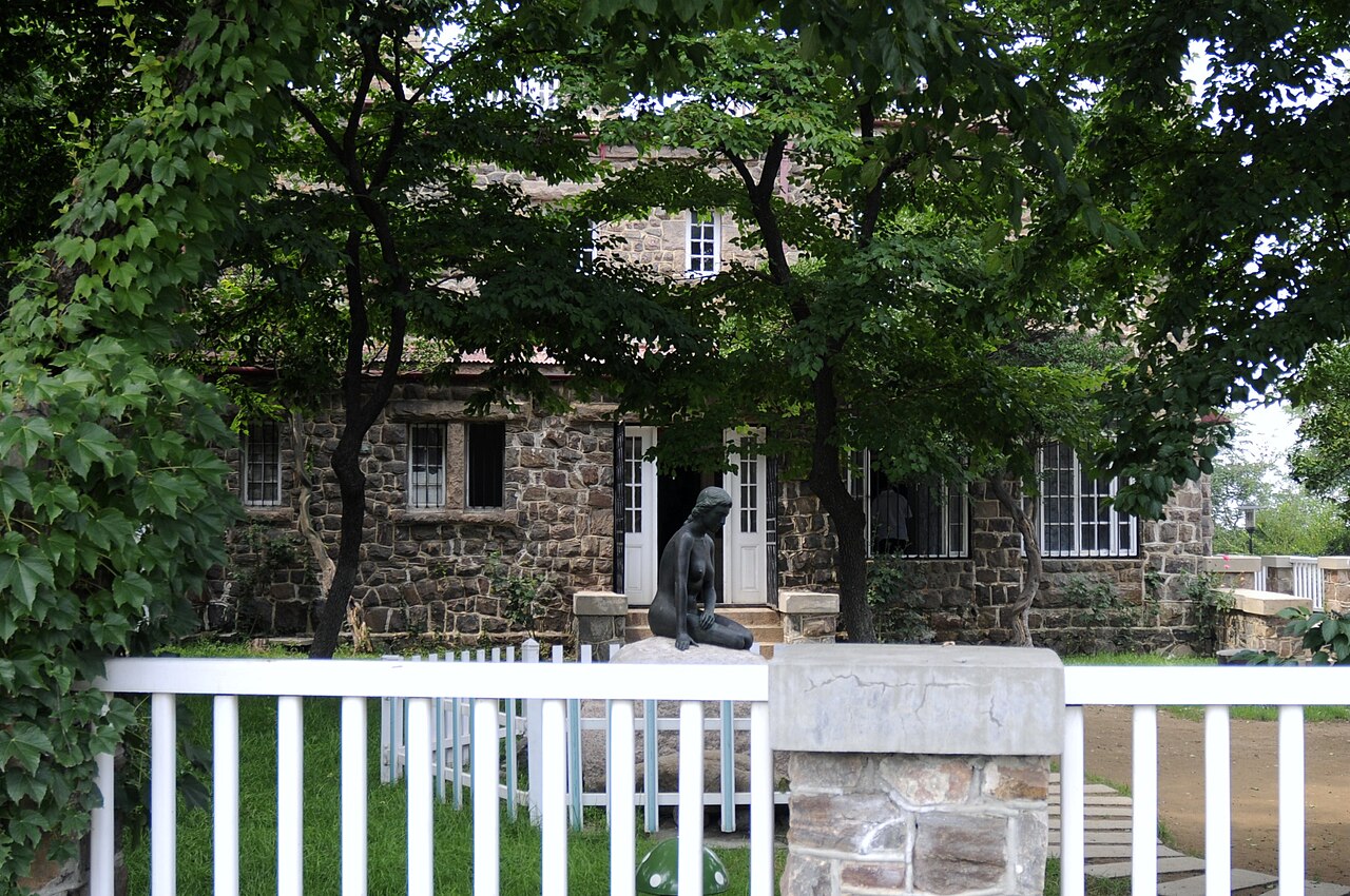 a grey stone house with tall windows behind a white fence and trees