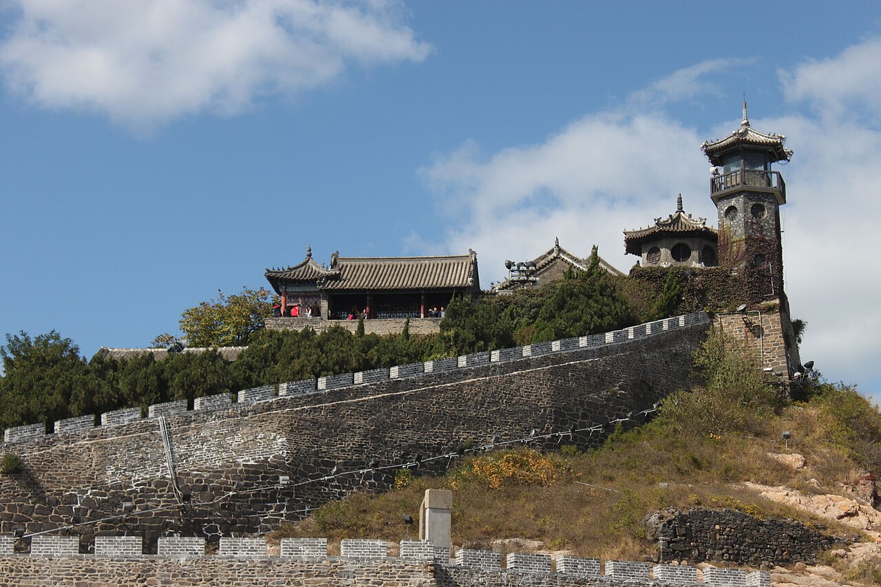 a tower and pavilions above an old crenellated wall on a grassy hillside under a blue sky