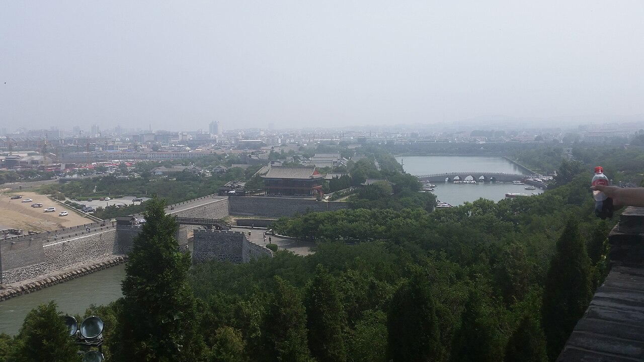 view from a hill over trees to a walled harbour basin and the town beyond