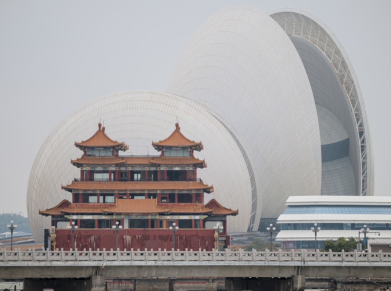 a red tiered Chinese building in front of a huge white shell-shaped roof