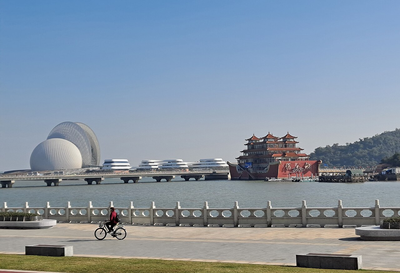 the white shells of the theatre and a red Chinese-style floating building seen across water from a seafront path with a cyclist